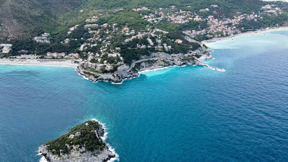 An aerial view of a coastal town with a prominent island, clear turquoise waters, and lush green hills in the background.