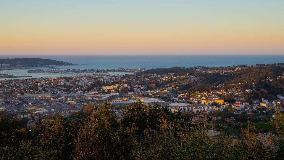 A coastal cityscape at dusk with a river, a bridge, and a hill with buildings.