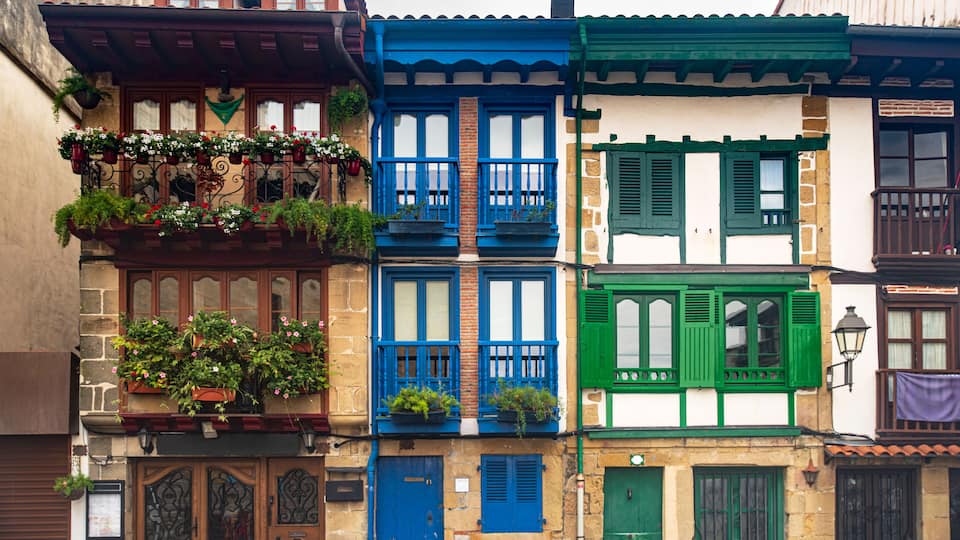 A row of colorful buildings with blue, green, and white facades, featuring balconies with plants and traditional shutters.