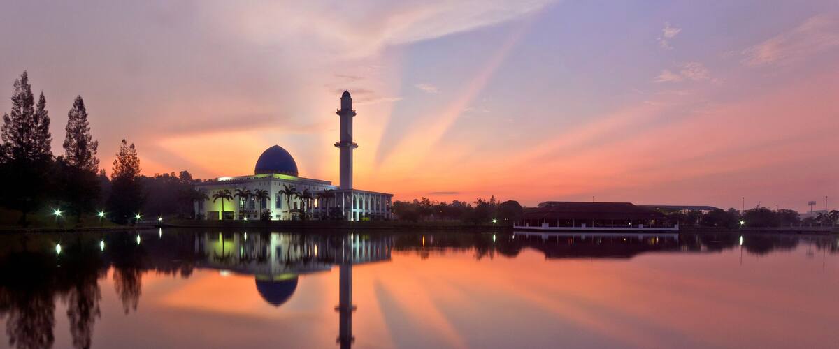 A mosque with a tall minaret and a dome, reflecting on a calm body of water during sunset.