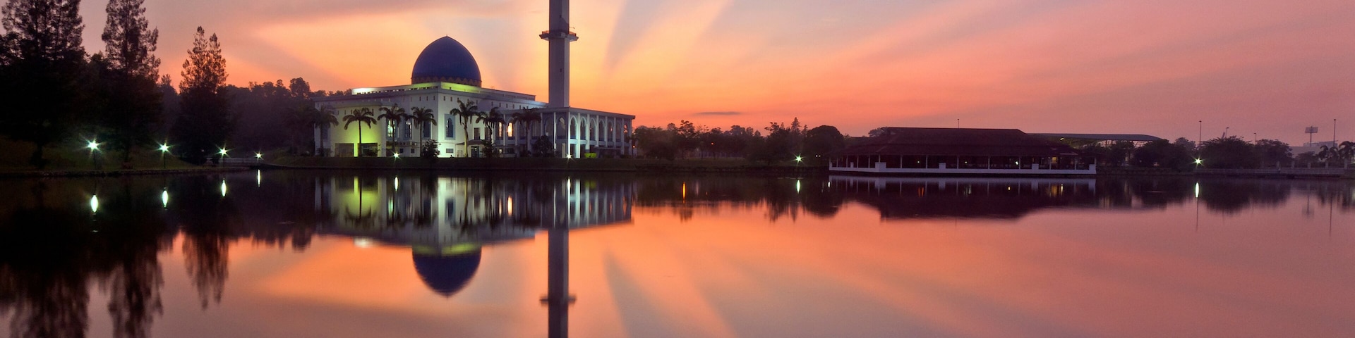 A mosque with a tall minaret and a dome, reflecting on a calm body of water during sunset.