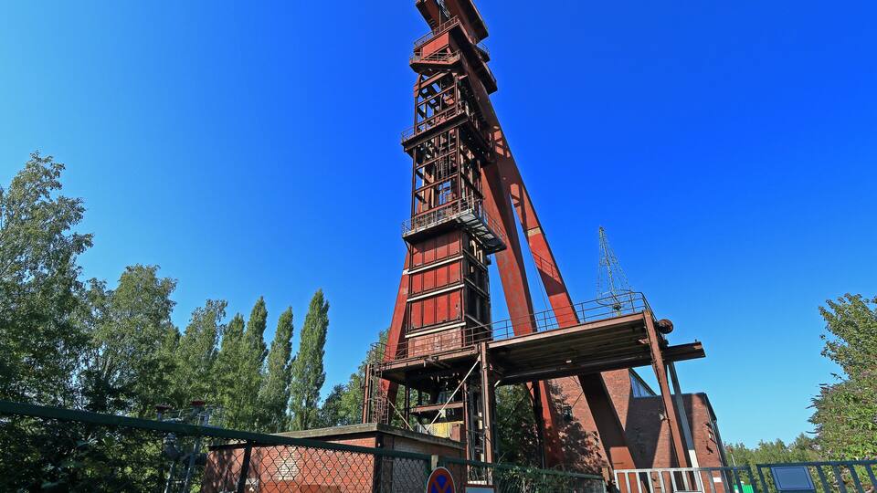 A large red industrial structure with a crane, surrounded by trees and a clear blue sky.