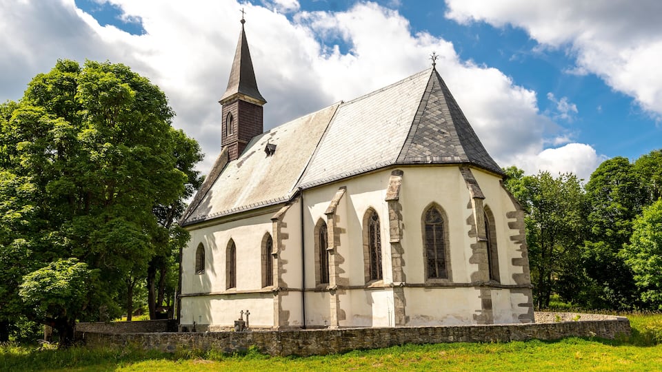 A church with a tall steeple and arched windows, surrounded by trees and a stone wall.