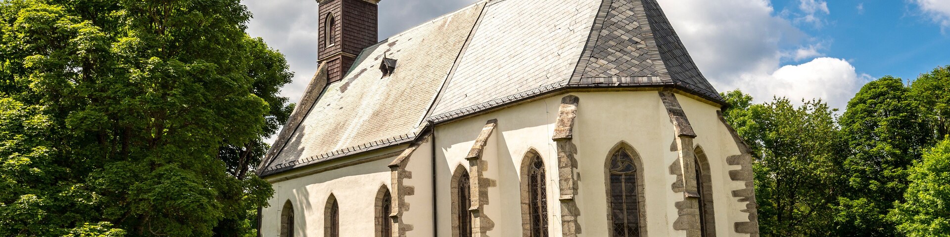 Una iglesia con un campanario alto y ventanas arqueadas, rodeada de árboles y un muro de piedra.