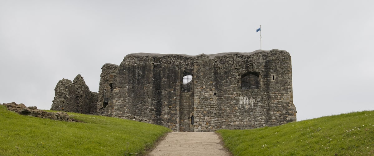 A stone castle with a flag on top, a pathway leading to the entrance, and grassy hills on either side.