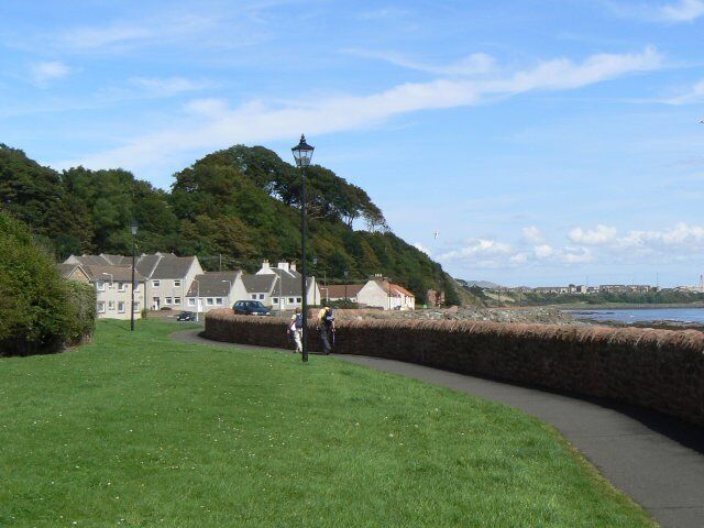 A coastal promenade with a grassy path, a stone wall, and houses in the background.
