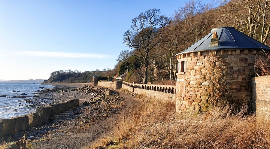A coastal stone wall with a turret, overlooking a rocky shoreline and a clear sky.