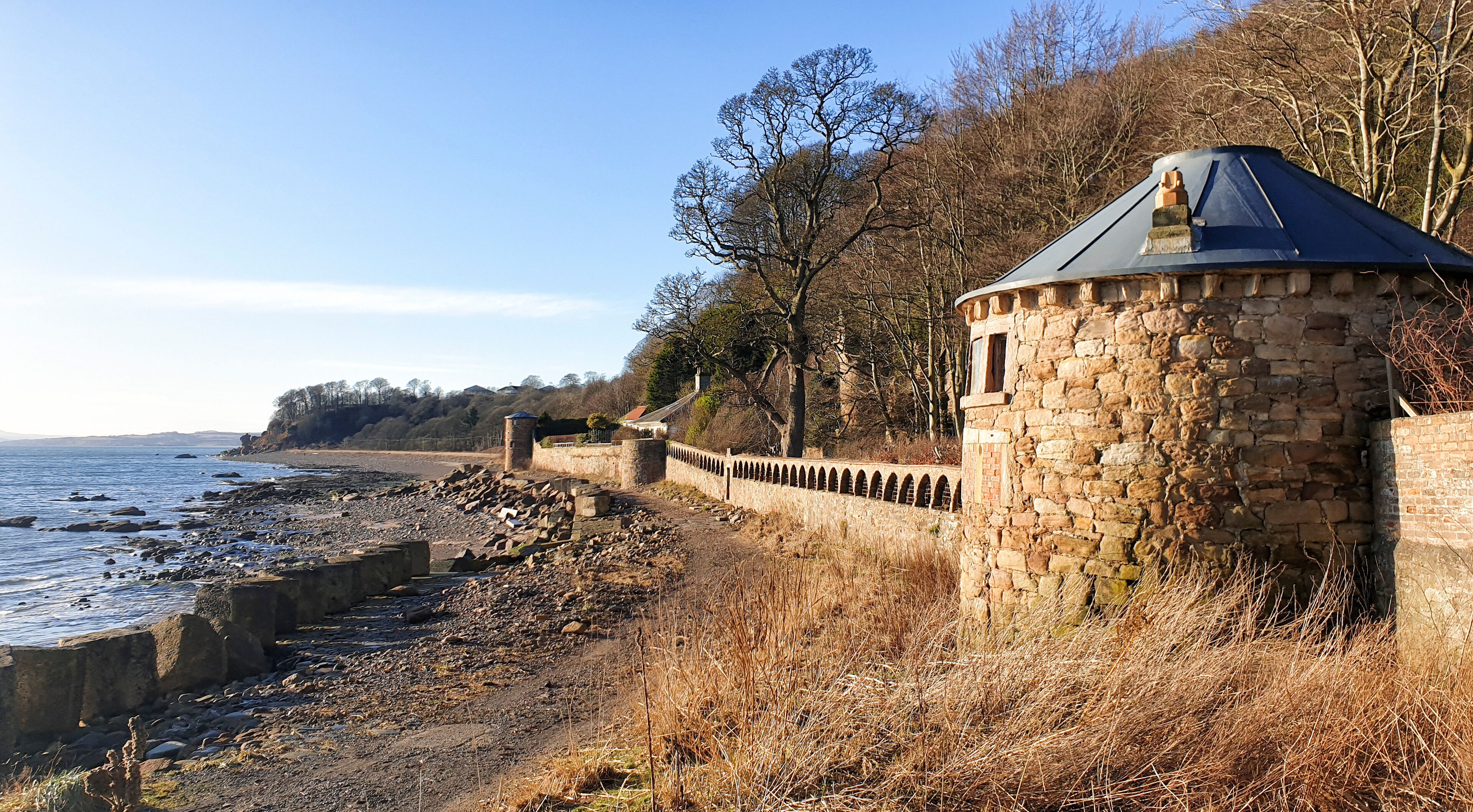 A coastal stone wall with a turret, overlooking a rocky shoreline and a clear sky.