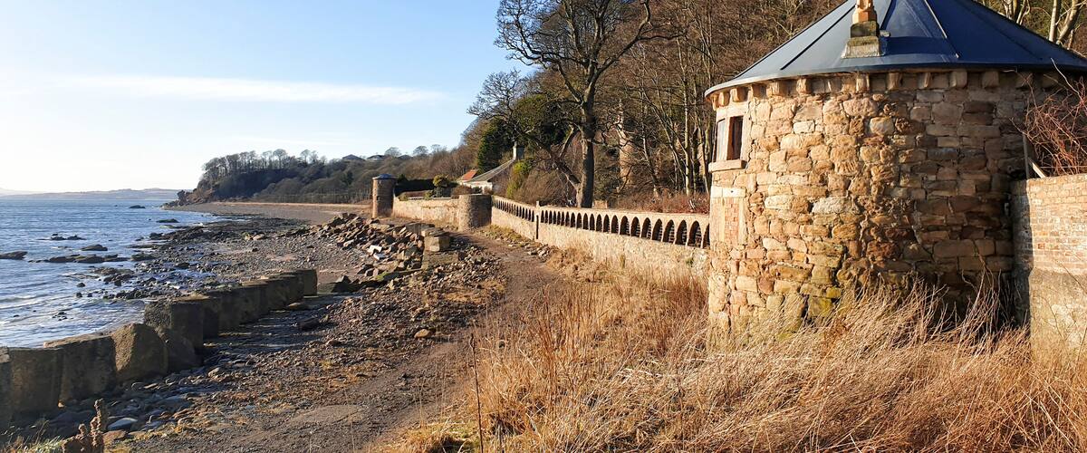 A coastal stone wall with a turret, overlooking a rocky shoreline and a clear sky.