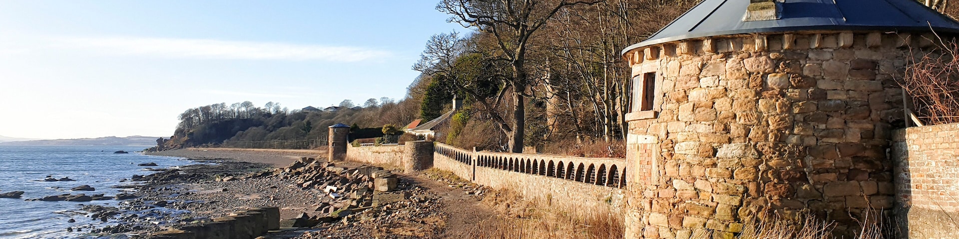A coastal stone wall with a turret, overlooking a rocky shoreline and a clear sky.