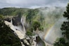 Une cascade se jetant en contrebas d’une falaise abrupte, entourée d’une végétation luxuriante et illuminée par un arc-en-ciel bien visible.