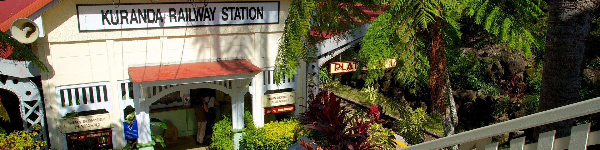 A railway station named Kuranda Railway Station with a red roof, surrounded by lush greenery and ferns.