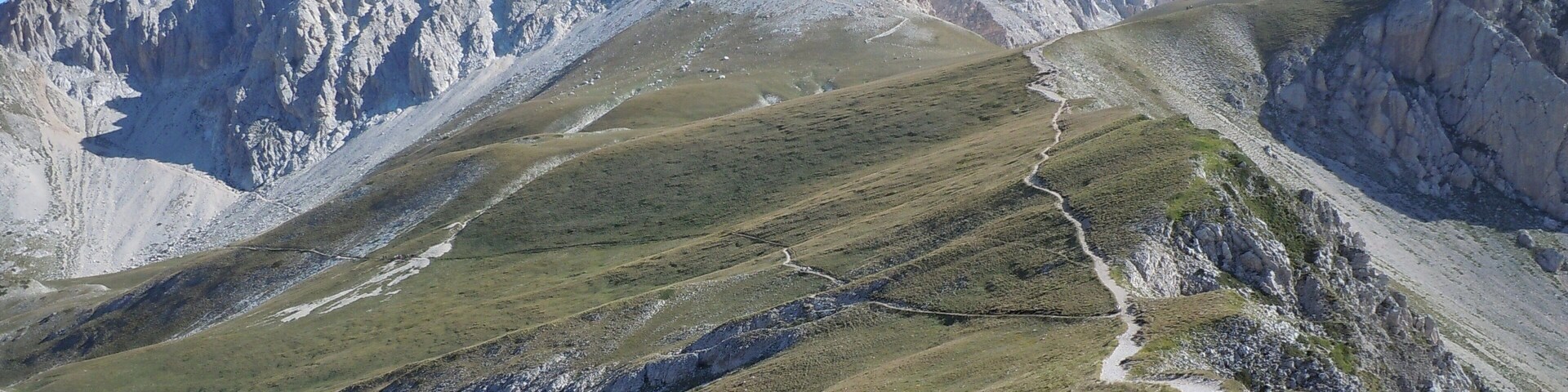 Paisagem montanhosa com pico rochoso, trilha sinuosa e vegetação rasteira.