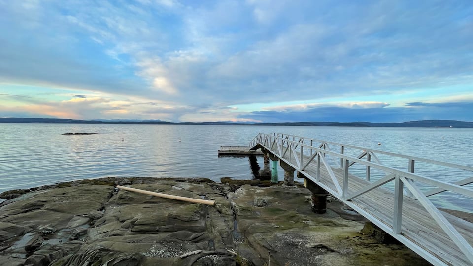 A wooden pier extending into calm waters, surrounded by rocky shores and distant mountains under a partly cloudy sky.