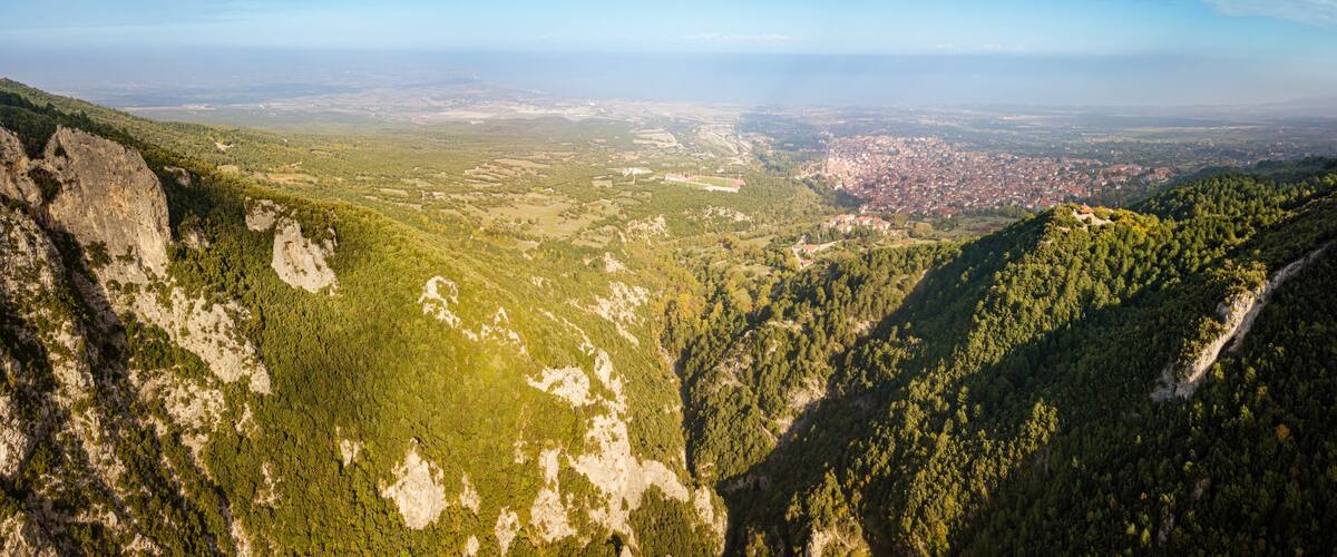 A mountainous landscape with dense forests, a valley, and a distant cityscape.