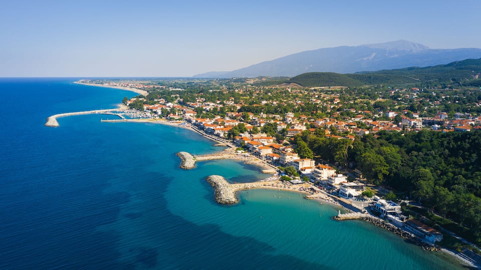 A coastal town with a clear blue sea, a winding harbor, and a mountainous backdrop.