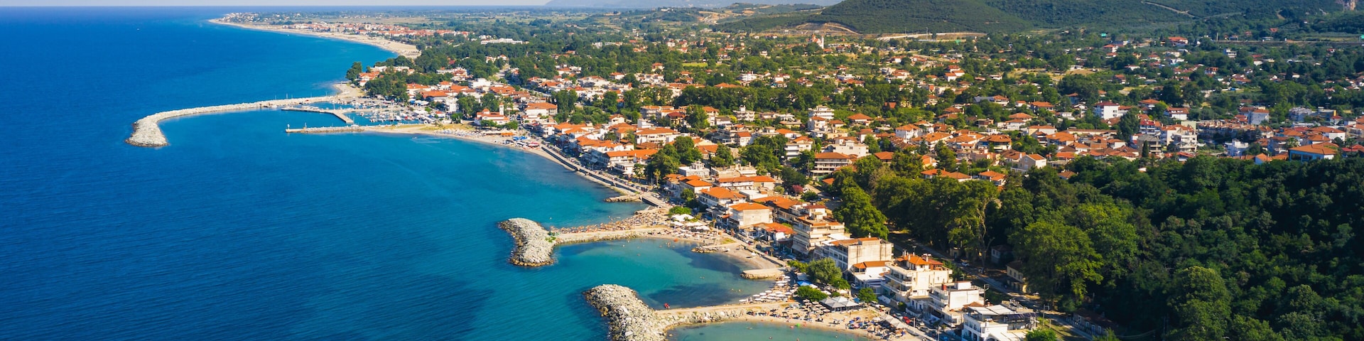 Un pueblo costero con mar azul cristalino, un puerto serpenteante y montañas al fondo.