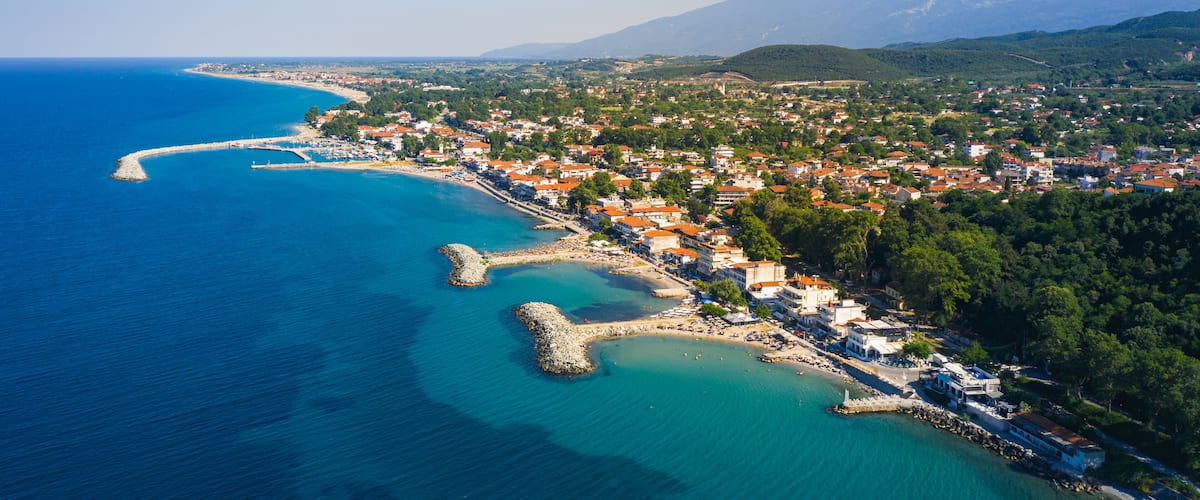 A coastal town with a clear blue sea, a winding harbor, and a mountainous backdrop.