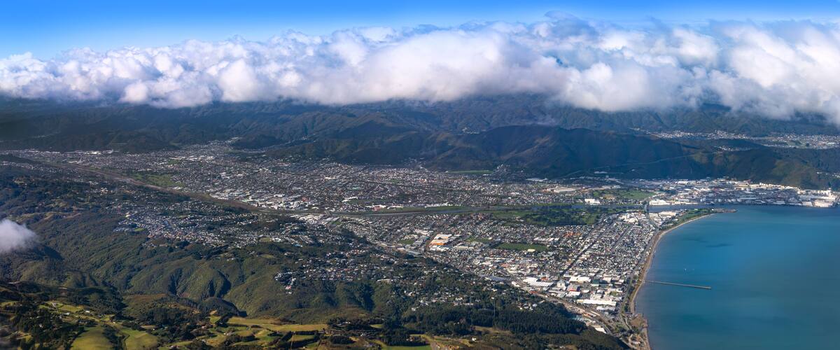 An aerial view of a coastal city with dense urban areas, a large body of water, and surrounding mountainous terrain partially covered by clouds.