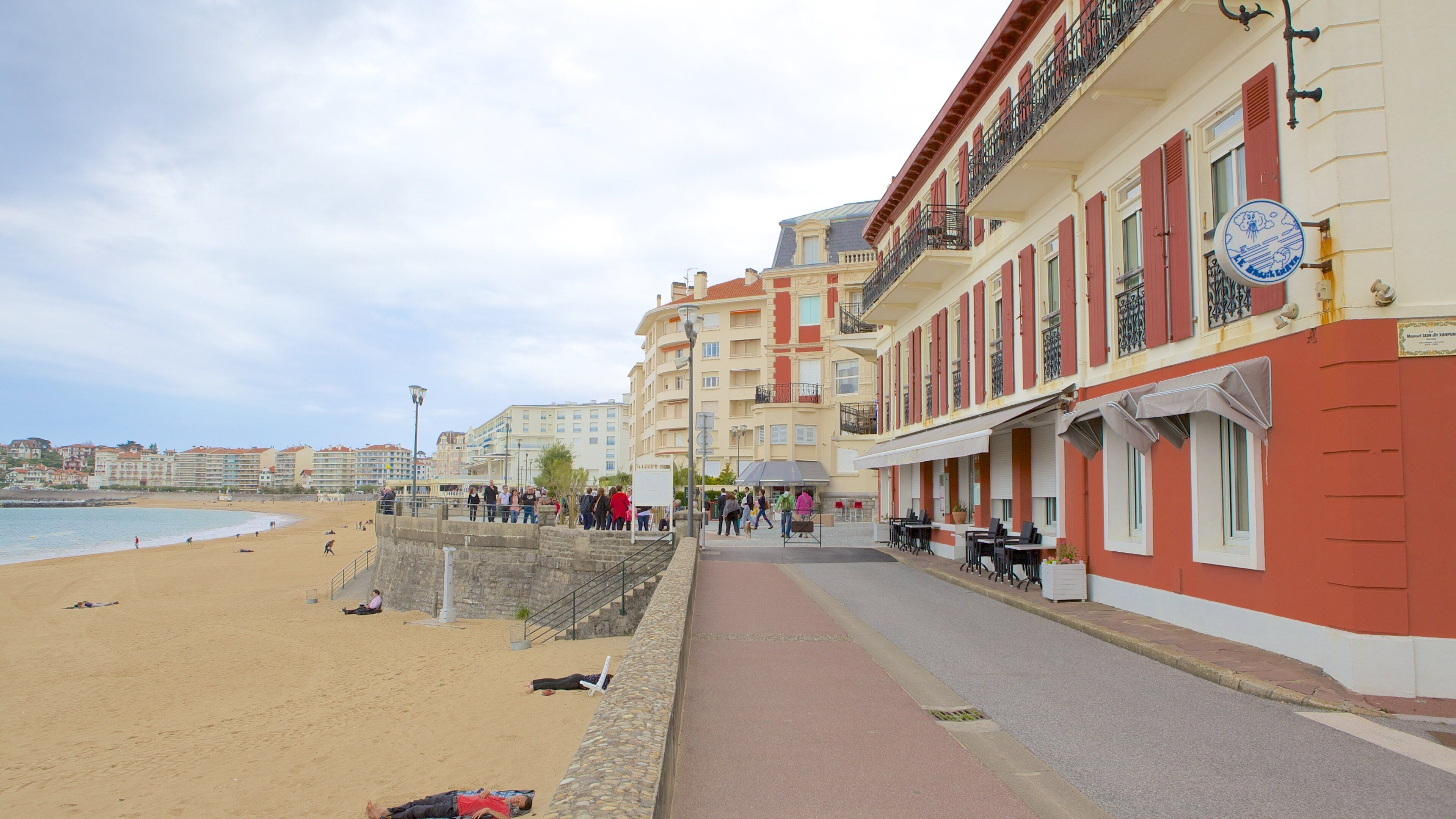 A beach with people sunbathing and walking along a promenade lined with buildings.