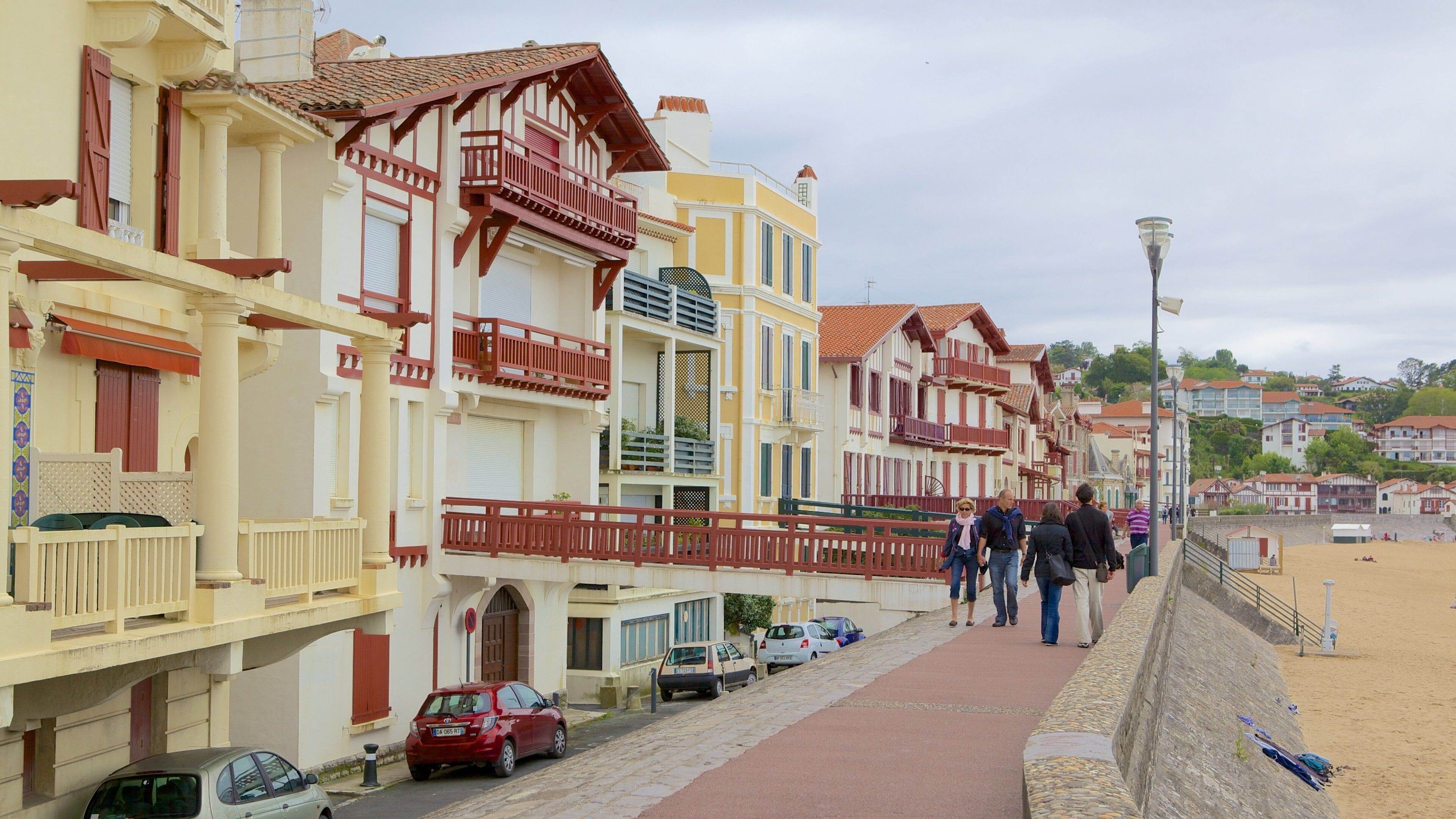 A coastal promenade with colorful buildings, a sandy beach, and parked cars.