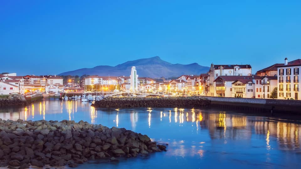 A coastal town at dusk with a mountain in the background, a bridge, and illuminated buildings reflecting on the water.