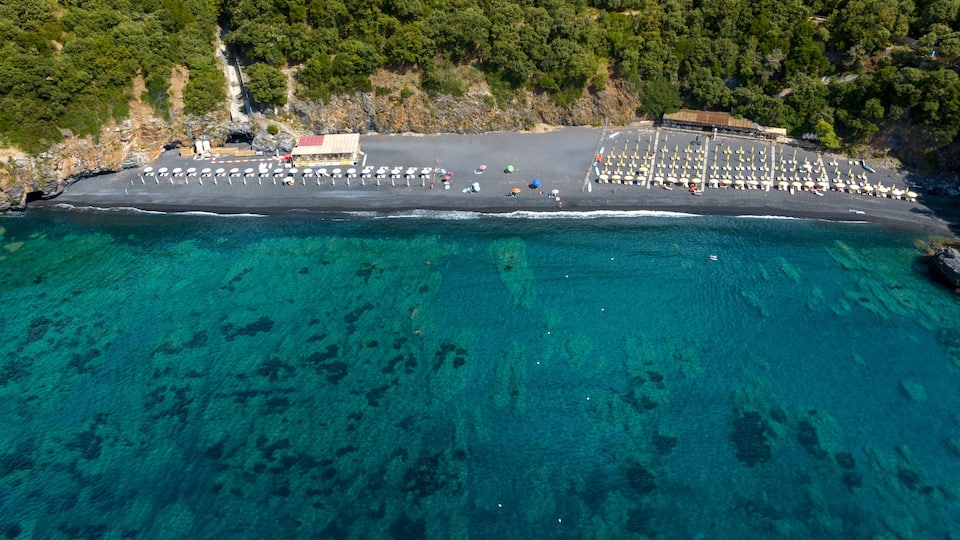 An aerial view of a beach with clear turquoise waters, a parking area with numerous umbrellas, and a building with a red roof.