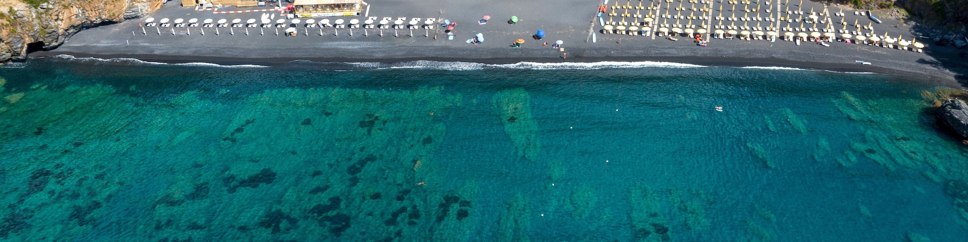 An aerial view of a beach with clear turquoise waters, a parking area with numerous umbrellas, and a building with a red roof.