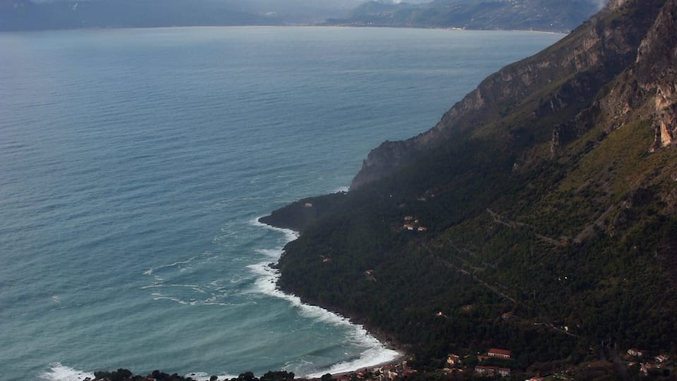 A coastal town nestled between cliffs, with a view of the sea and distant mountains.