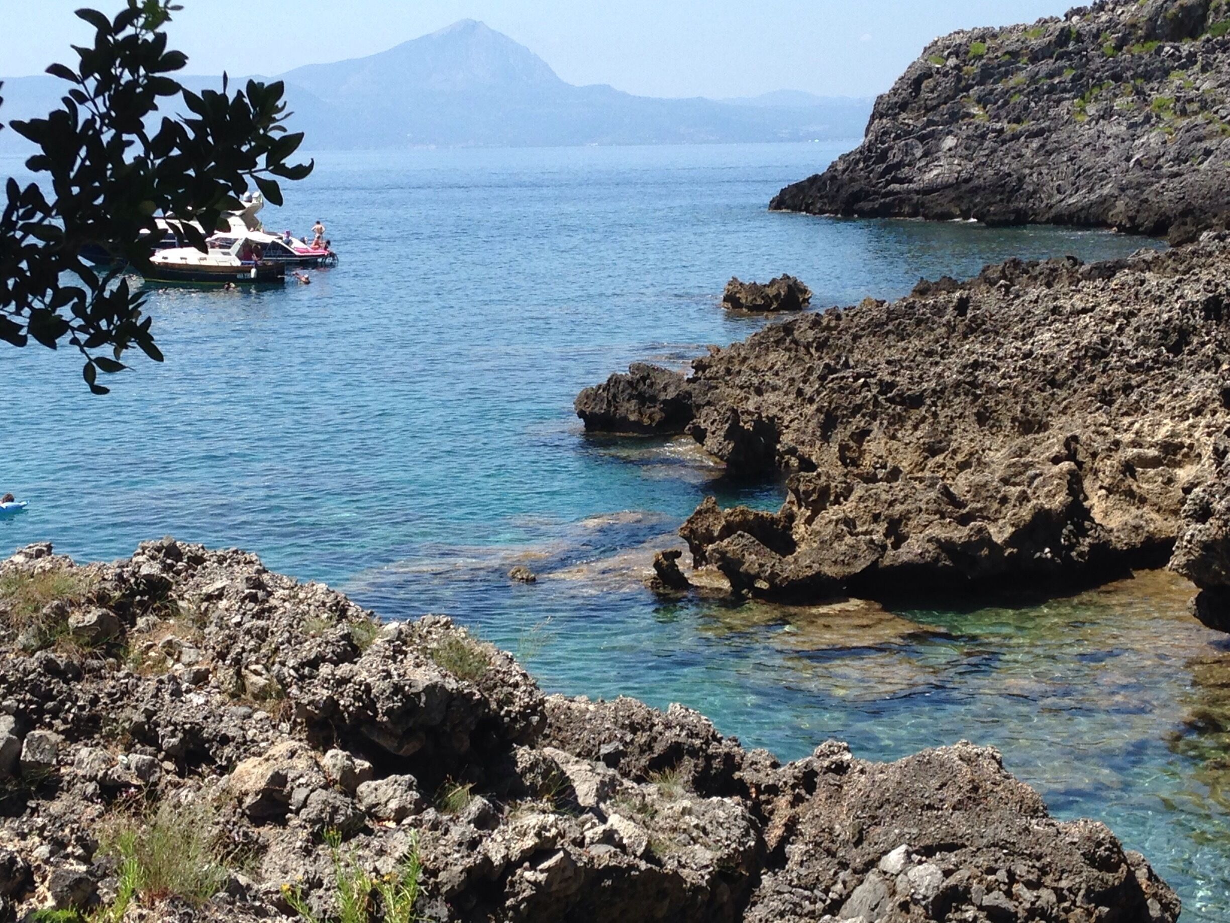 A rocky coastline with clear blue water, a boat in the distance, and a mountain in the background.