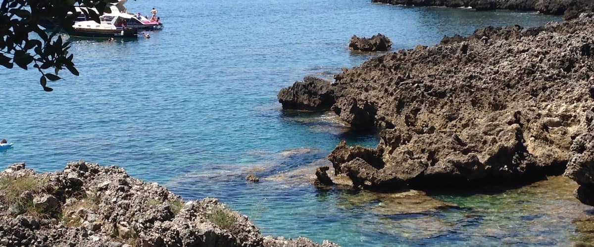 A rocky coastline with clear blue water, a boat in the distance, and a mountain in the background.