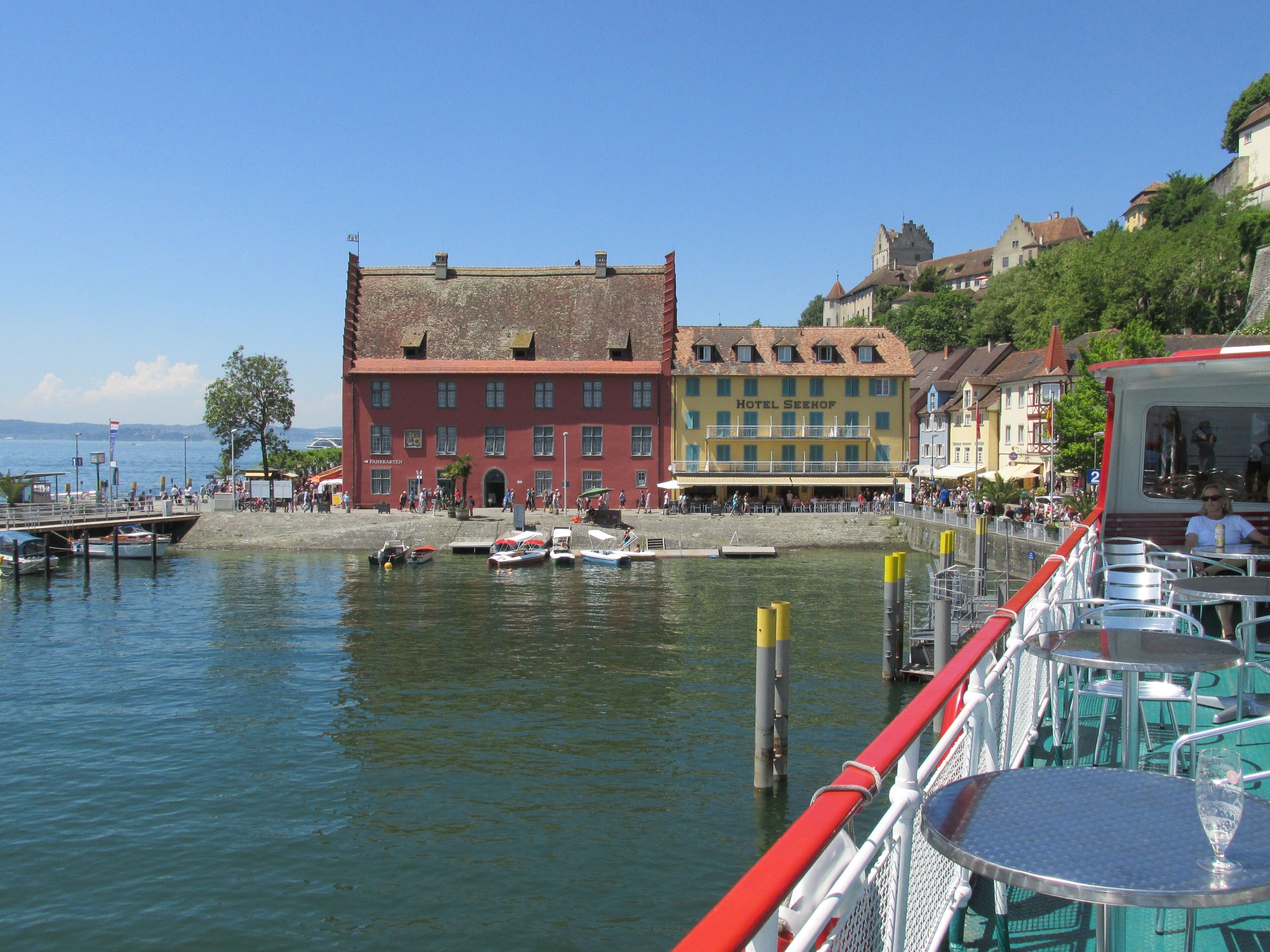 A waterfront with boats docked, a red building labeled "Hotel Seehof," and a castle on a hill.