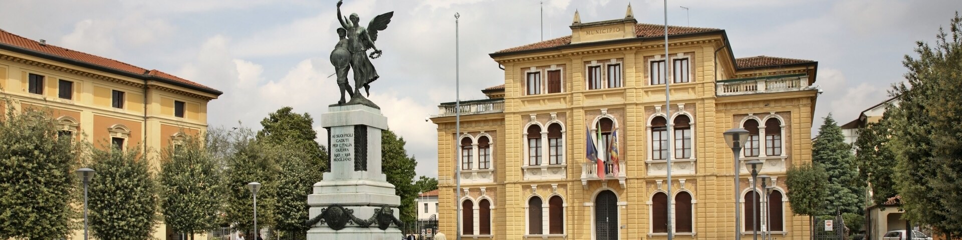 A square with a statue, a yellow building, and trees.