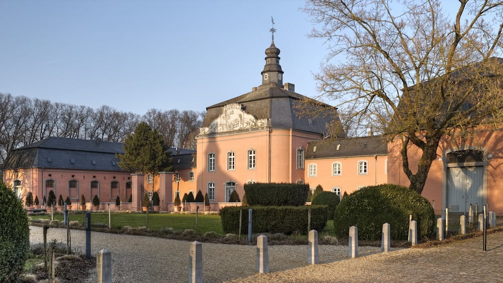 Ein großes, pinkfarbenes Gebäude mit einem Turm, umgeben von einem Kiesweg und geschnittenen Hecken.