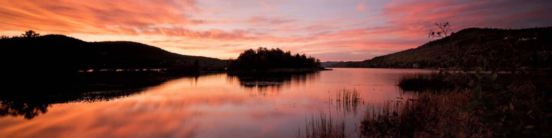 A calm lake at sunset with a reflection of the sky, surrounded by hills and vegetation.