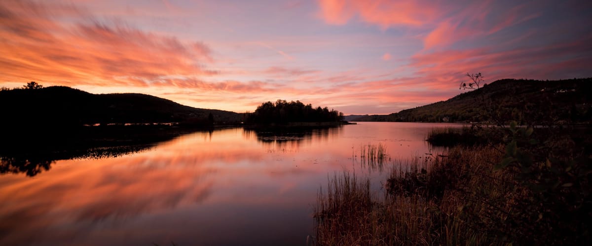 A calm lake at sunset with a reflection of the sky, surrounded by hills and vegetation.
