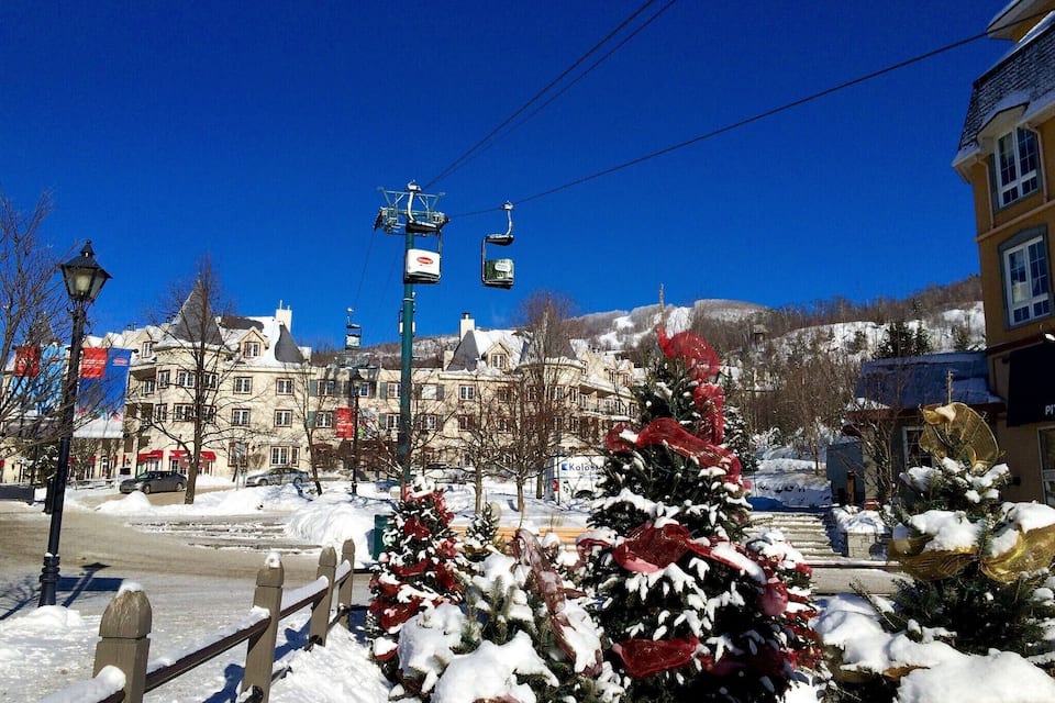 A snowy town square with a decorated Christmas tree, a ski lift, and buildings in the background.