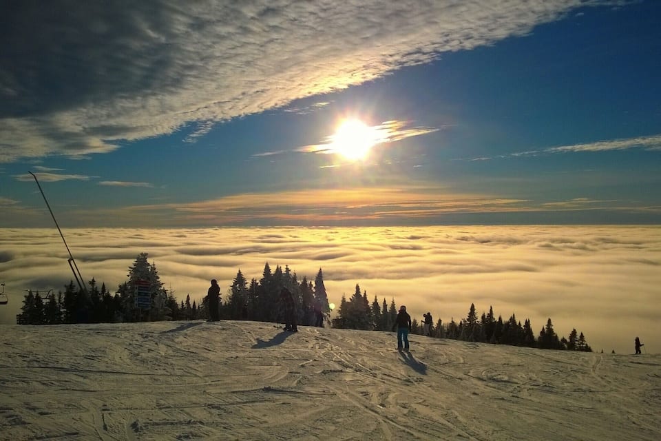 Paisaje nevado de montaña con un telesilla, árboles y un sol poniéndose tras las nubes.