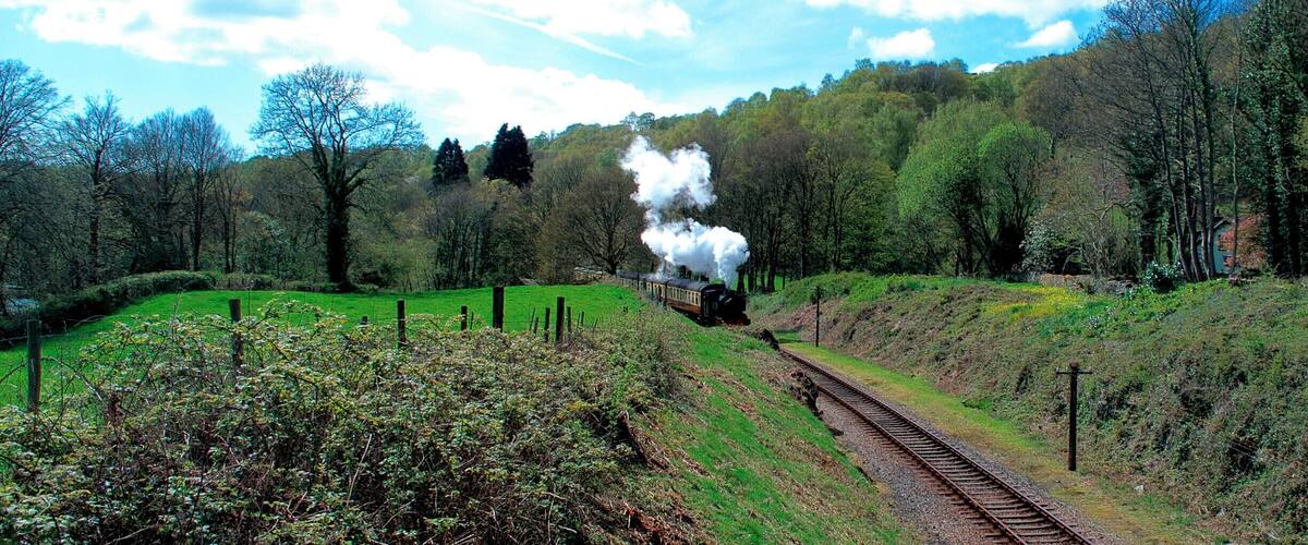 A train with white smoke on a track through a green landscape with trees and hills.