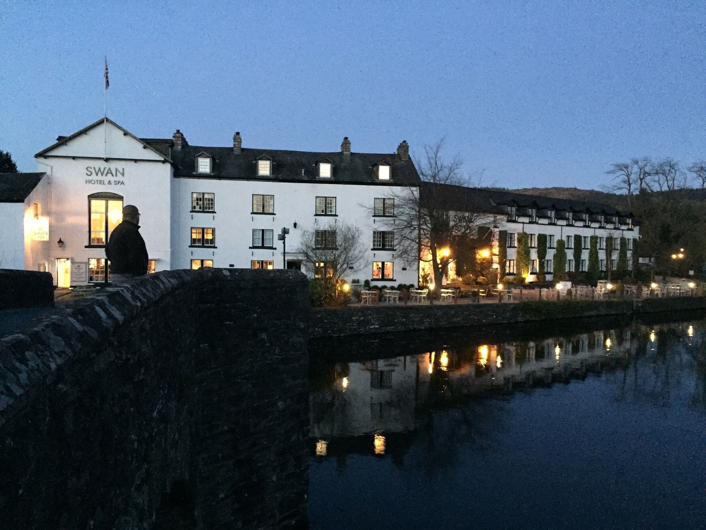 A white building with a sign reading "SWAN HOTEL & SPA" by a calm river.
