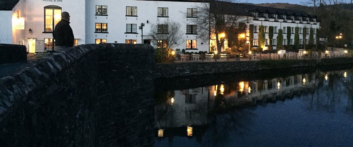 A white building with a sign reading "SWAN HOTEL & SPA" by a calm river.