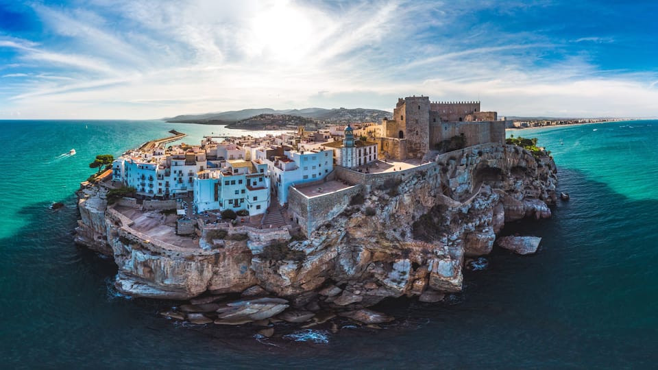 A coastal town with a prominent castle on a rocky outcrop, surrounded by turquoise waters and clear skies.