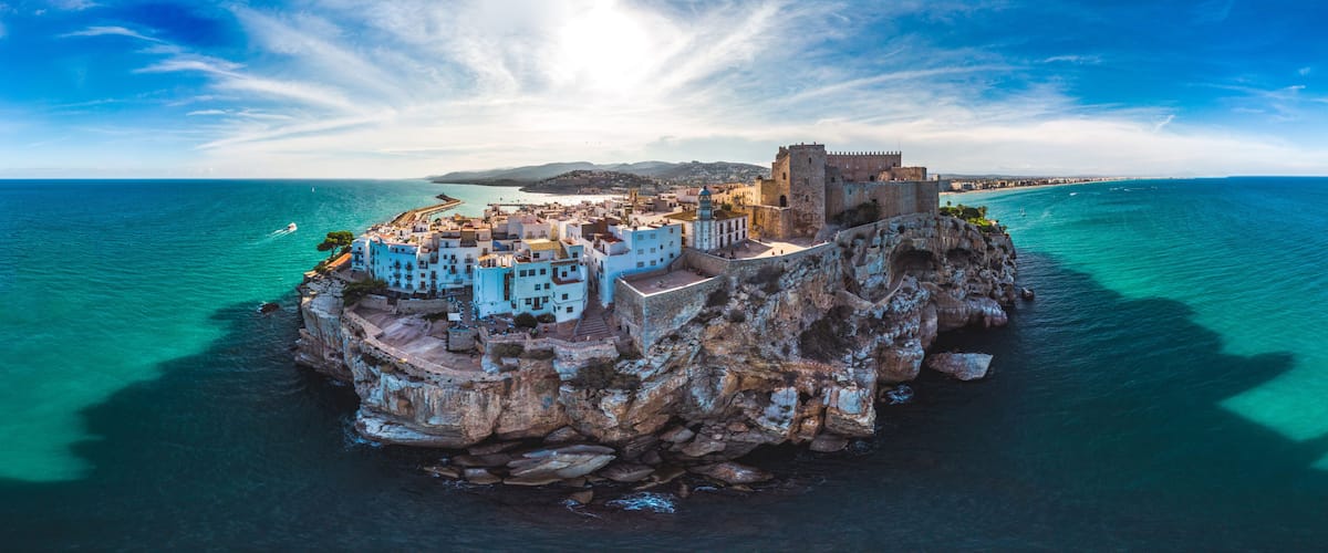 A coastal town with a prominent castle on a rocky outcrop, surrounded by turquoise waters and clear skies.