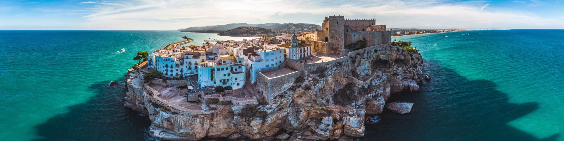 A coastal town with a prominent castle on a rocky outcrop, surrounded by turquoise waters and clear skies.