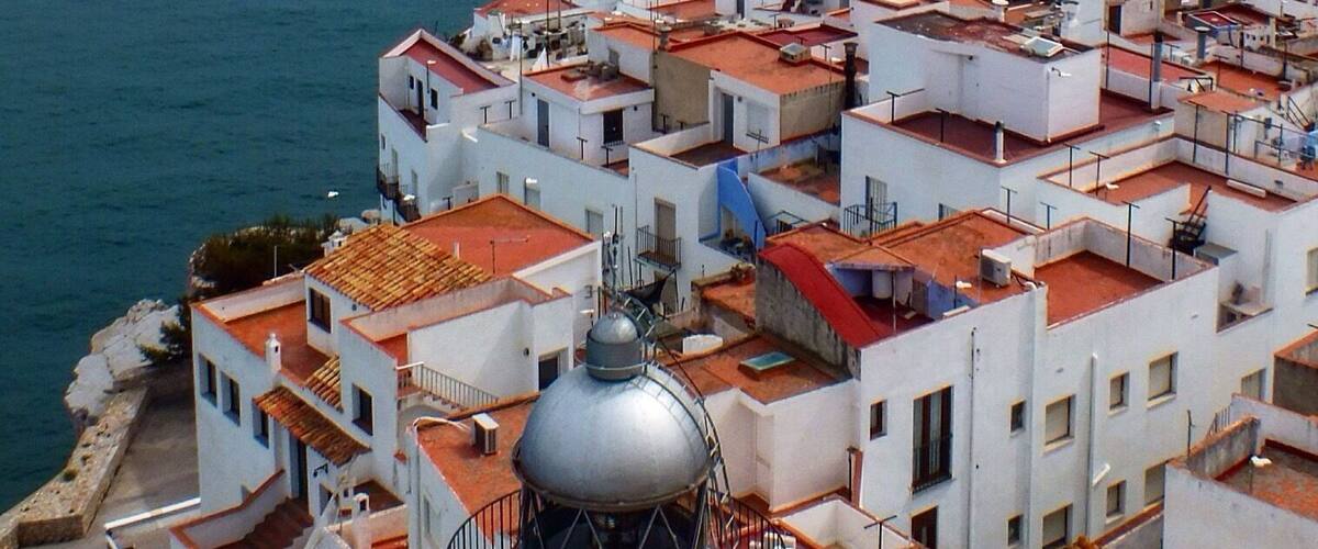 Un pueblo costero con edificios blancos y tejados rojos, una estructura en forma de cúpula destacada, y un barco en el agua.