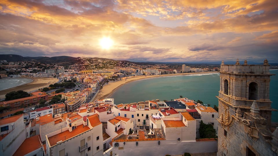 A coastal cityscape with a prominent tower, a beach, and a mountain range in the background.