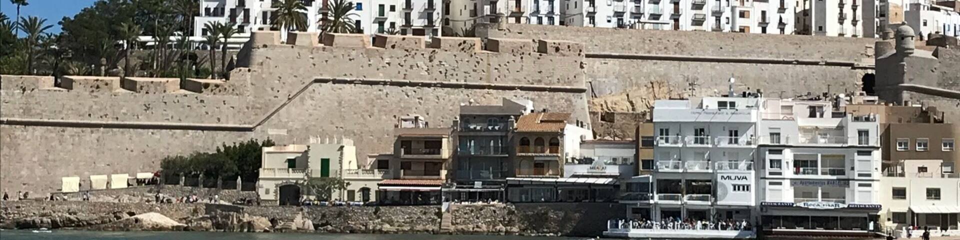 A coastal city with a prominent fortress on a hill, sandy beaches, and a clear blue sky.