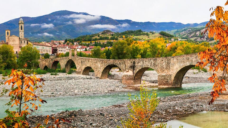 A stone bridge over a river, surrounded by autumn foliage and a mountainous backdrop.