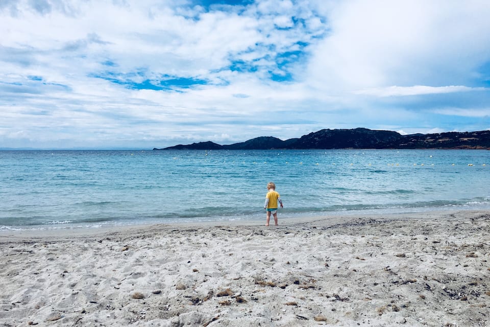 A beach with a child in a yellow shirt standing on the sand, facing the sea.