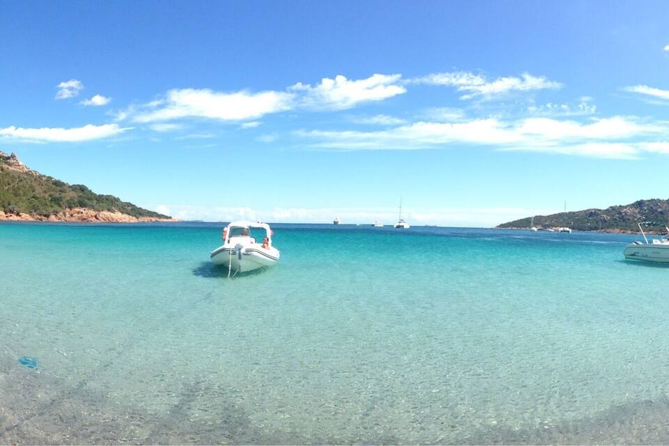 Une mer d’un bleu turquoise limpide, où des bateaux sont amarrés près du rivage, le tout entouré de collines et d’une végétation clairsemée.
